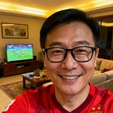 一位40岁左右戴眼镜的中国男性球迷头像，身穿红色球衣，面带微笑，背景为家中客厅观赛场景